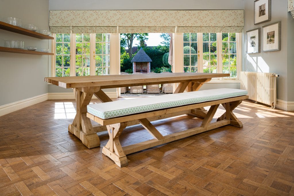 A large wooden dining table with cushioned benches in a dining room upon a wooden parqueted flooring