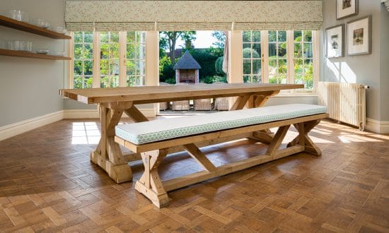 A large wooden dining table with cushioned benches in a dining room upon a wooden parqueted flooring