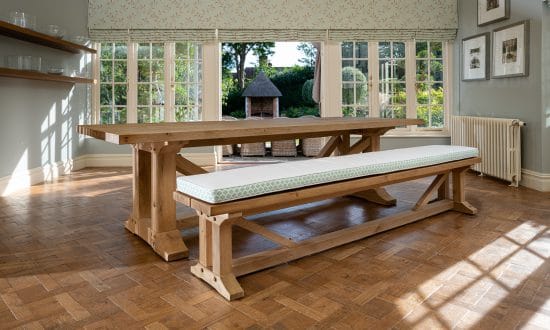 A large wooden dining table with cushioned bench seating in a dining room upon a wooden parqueted flooring