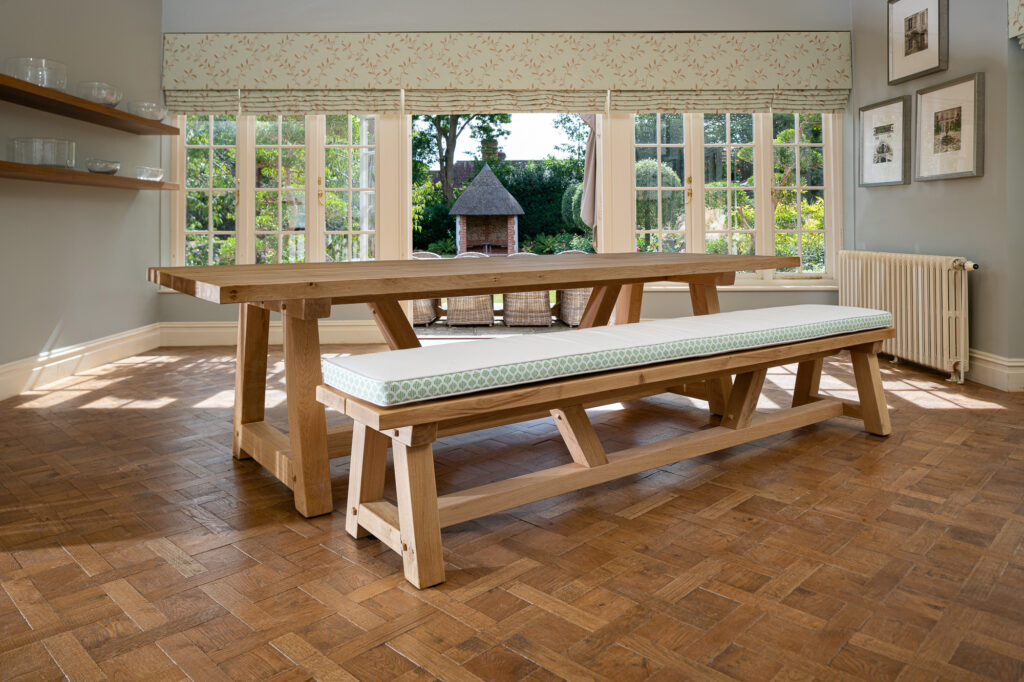 Oxenwood's wooden hollom large format dining table with cushioned benches shown in a parquet floored dining room